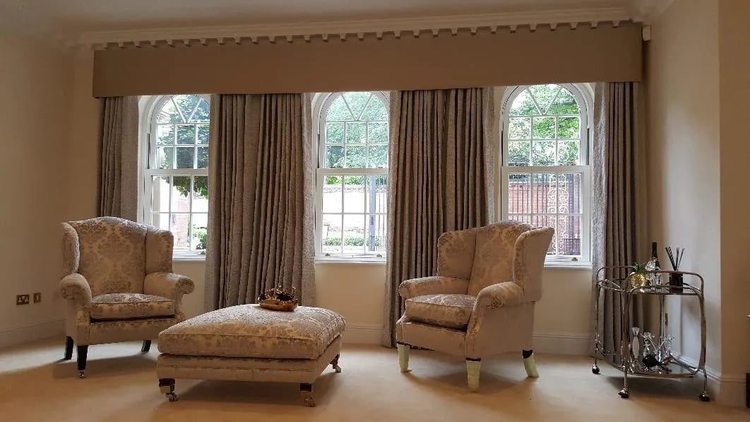 Arched Gothic windows dressed with a shaped and trimmed pelmet running the full width, and interlined curtains held back to either side. Period glazing like this needs the pelmet drawn to the arch, not squared off above it.