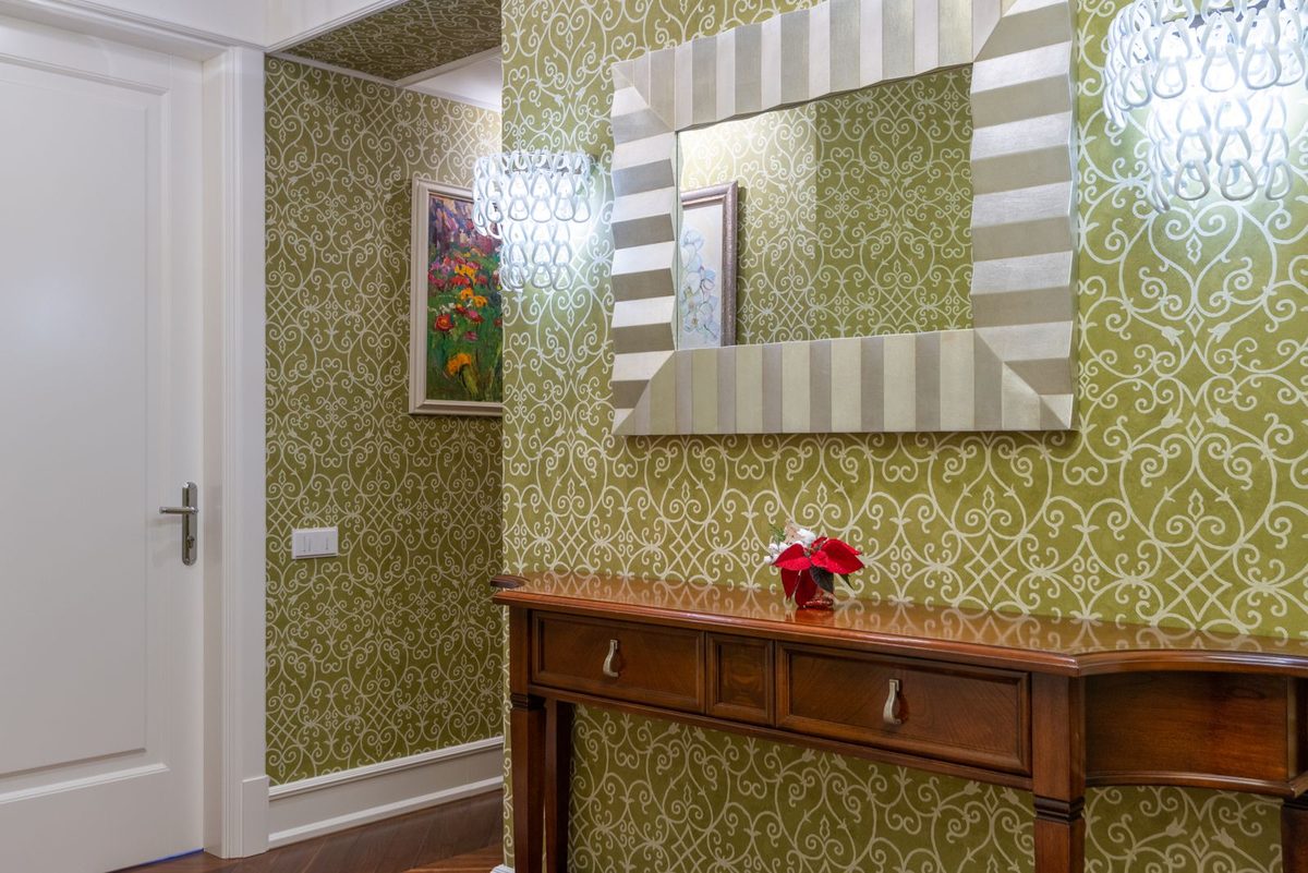 A hallway papered in a patterned wallpaper with a console table and mirror